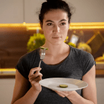 Woman holding plate with cucumber slice in kitchen, contemplating a healthy meal choice.