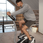 Man exercising on a stationary bike in a home gym with large windows in the background